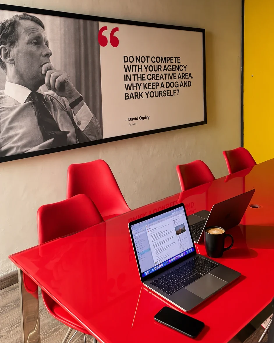 A meeting room with red chairs, an open laptop and a coffee mug beneath a framed David Ogilvy quote: “Do not compete with your agency in the creative area. Why keep a dog and bark yourself?”